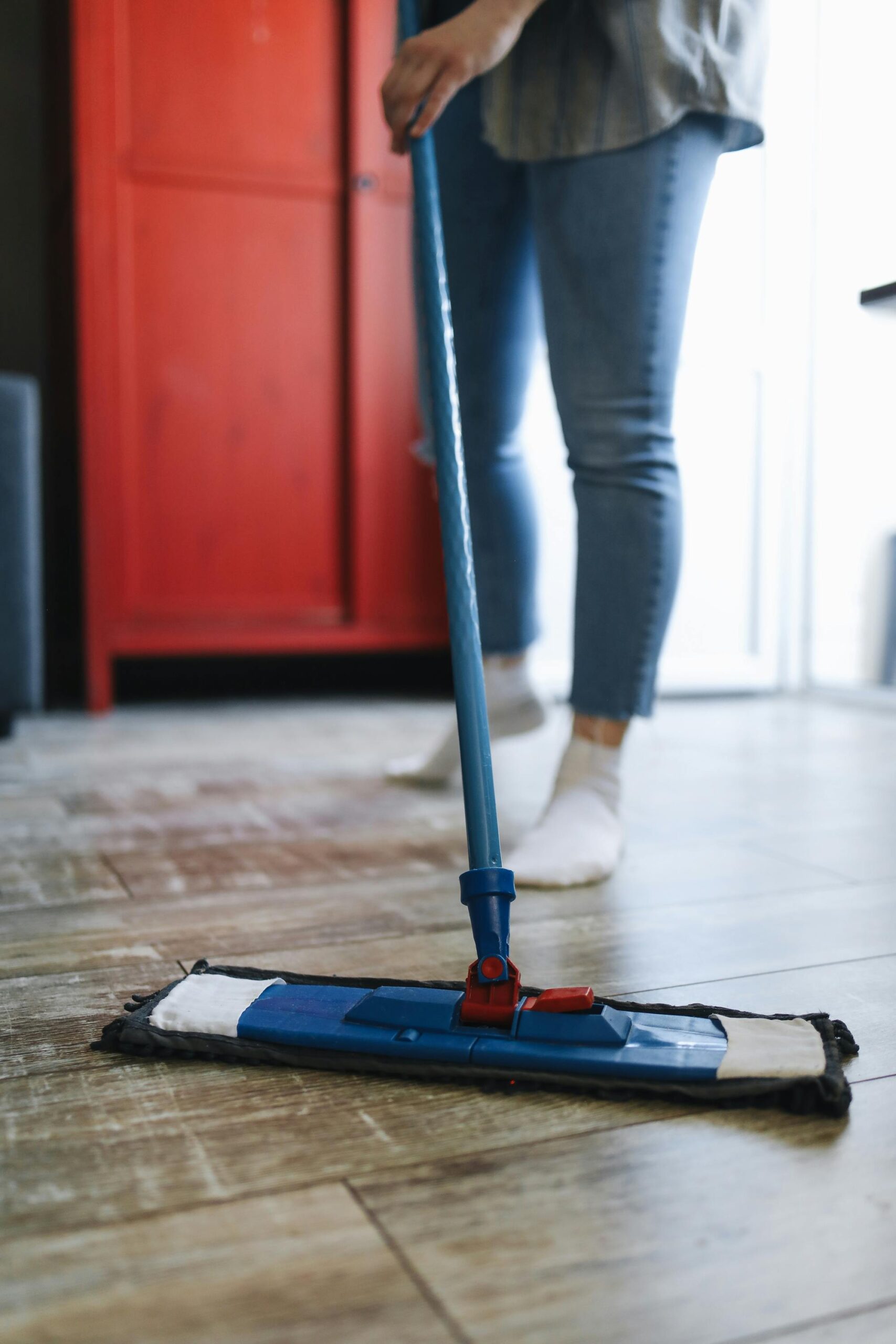 A woman mops a wooden floor in a modern living room with a red cabinet, emphasizing cleanliness.